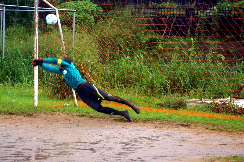 A goalkeeper in teal diving at full stretch as the ball flies past the post